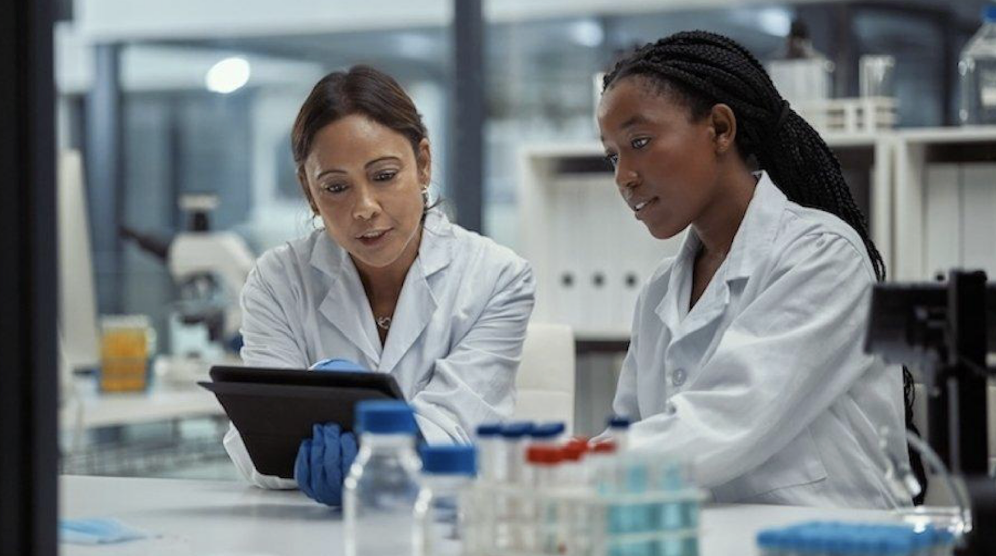 Two female scientists in lab coats examining data on a tablet